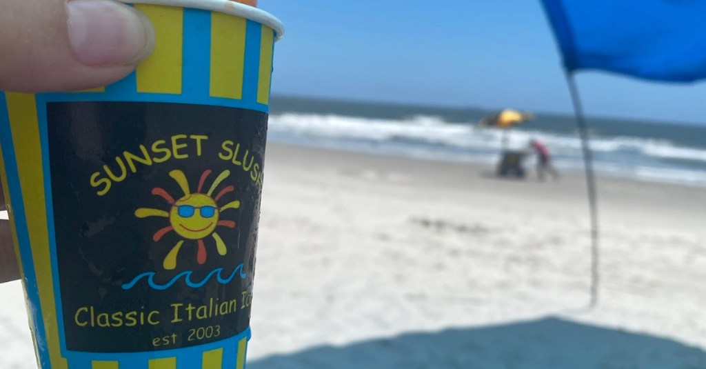 Sunset Slush Italian Ice cup held up in front of Holden Beach shoreline